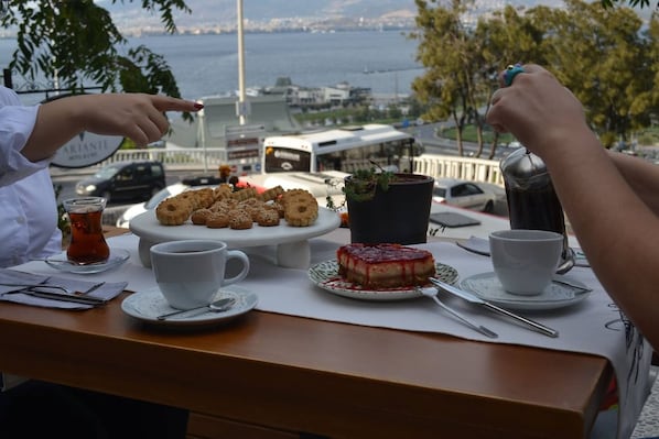 A table with a variety of pastries, a plate with a dessert, and a cup of tea, set against a backdrop of a cityscape and water.