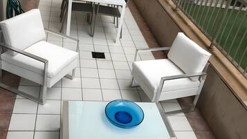 Een balkon met een witte tafel, twee witte stoelen, een blauwe schaal en uitzicht op een gebouw.