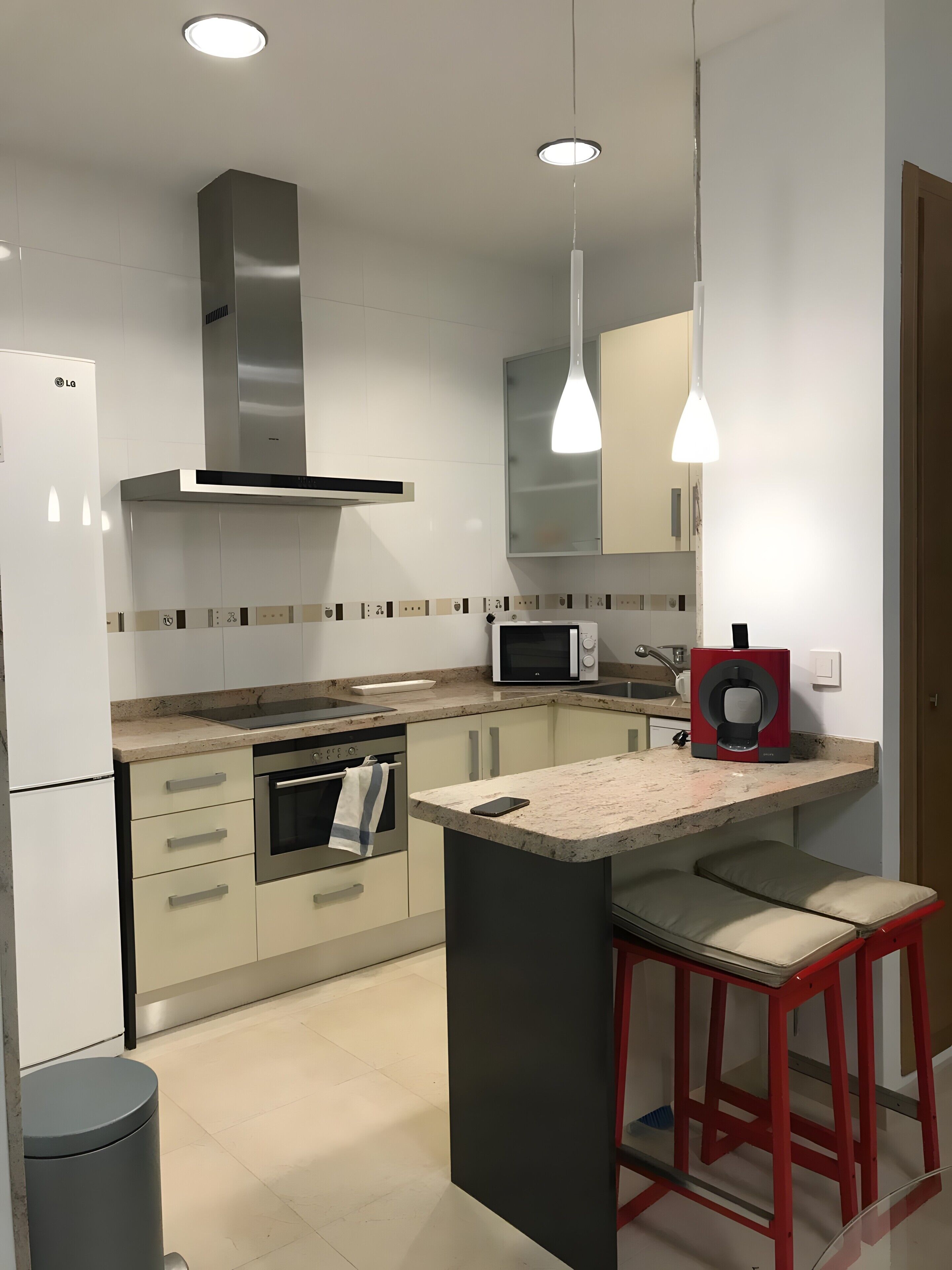 Een moderne keuken met een magnetron, oven en een koffieapparaat. Er staat een barstoel en een afvalbak.