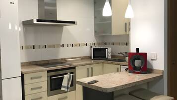 Een moderne keuken met een magnetron, oven en een koffieapparaat. Er staat een barstoel en een afvalbak.