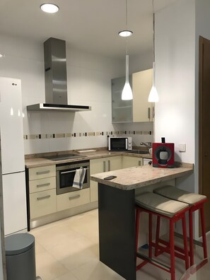 Eine moderne Küche mit Mikrowelle, Backofen und Kaffeemaschine. Ein Barhocker und ein Mülleimer sind vorhanden.