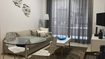 Een moderne woonkamer met een glazen eettafel, een bank, een televisie en een groot raam met gordijnen.