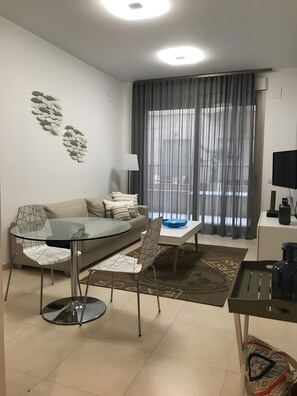 Ein modernes Wohnzimmer mit einem Glastisch, einer Couch, einem Fernseher und einem großen Fenster mit Vorhängen.