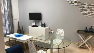 Een moderne woonkamer met een glazen eettafel, twee stoelen, een aan de muur bevestigde televisie en een decoratief kunstwerk van vissen.