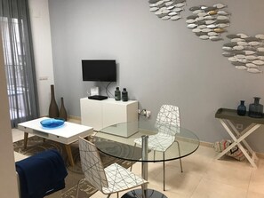 Ein modernes Wohnzimmer mit einem Glastisch, zwei Stühlen, einem an der Wand montierten Fernseher und einem dekorativen Wandbild von Fischen.