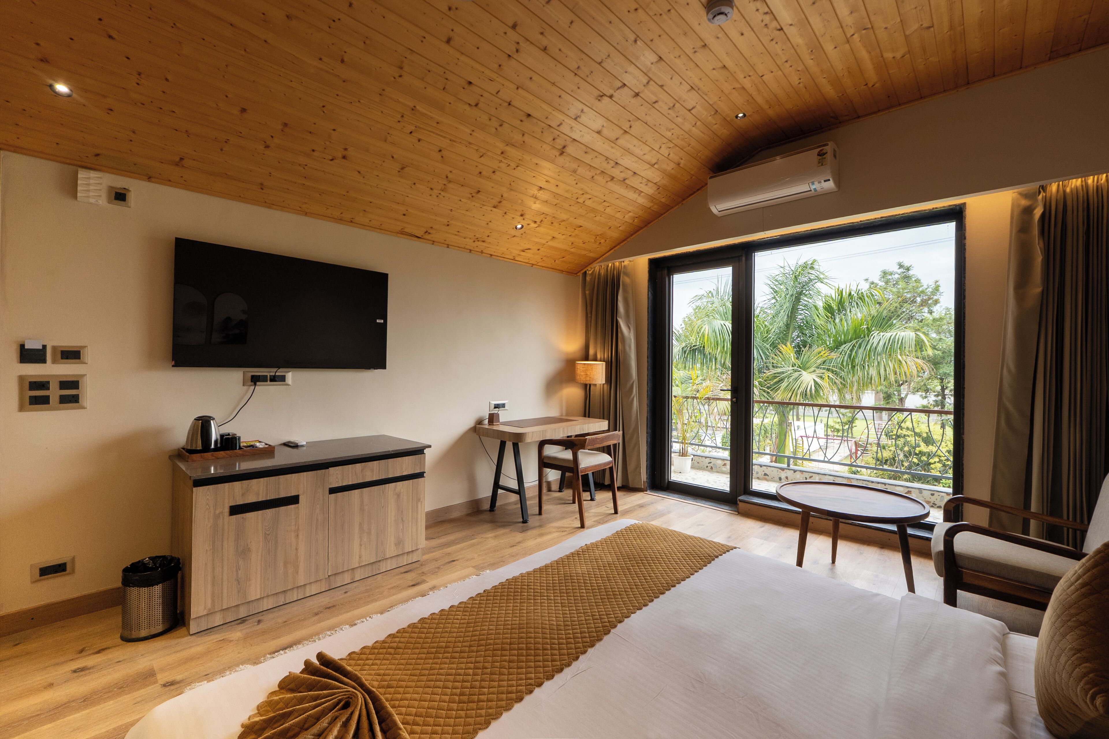 Habitación de hotel con techo de madera, televisor montado en la pared, cama con manta marrón, escritorio con silla y balcón con vistas a palmeras.