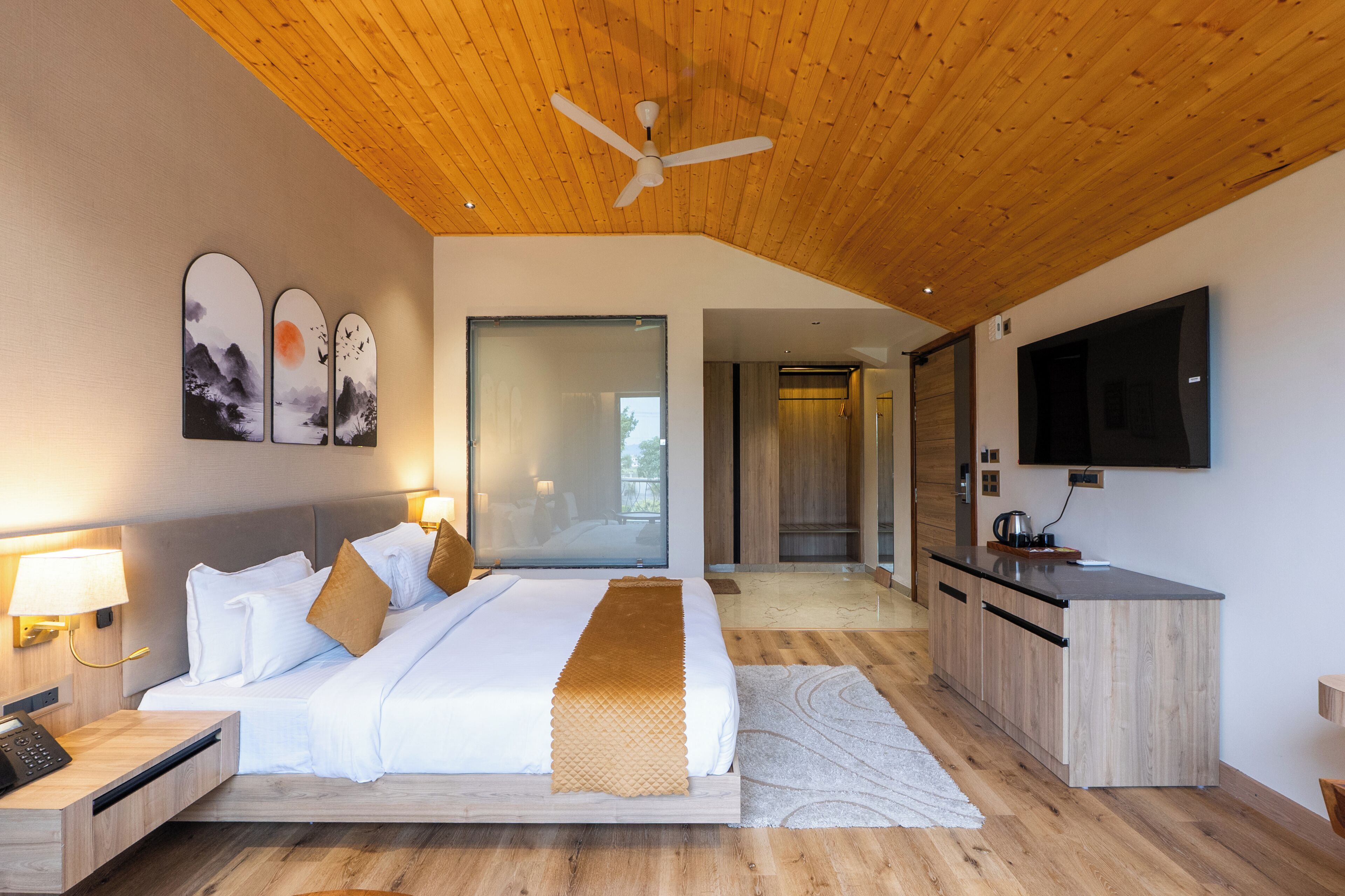 A modern hotel room with a bed, bedside table, lamp, artwork, and a TV.