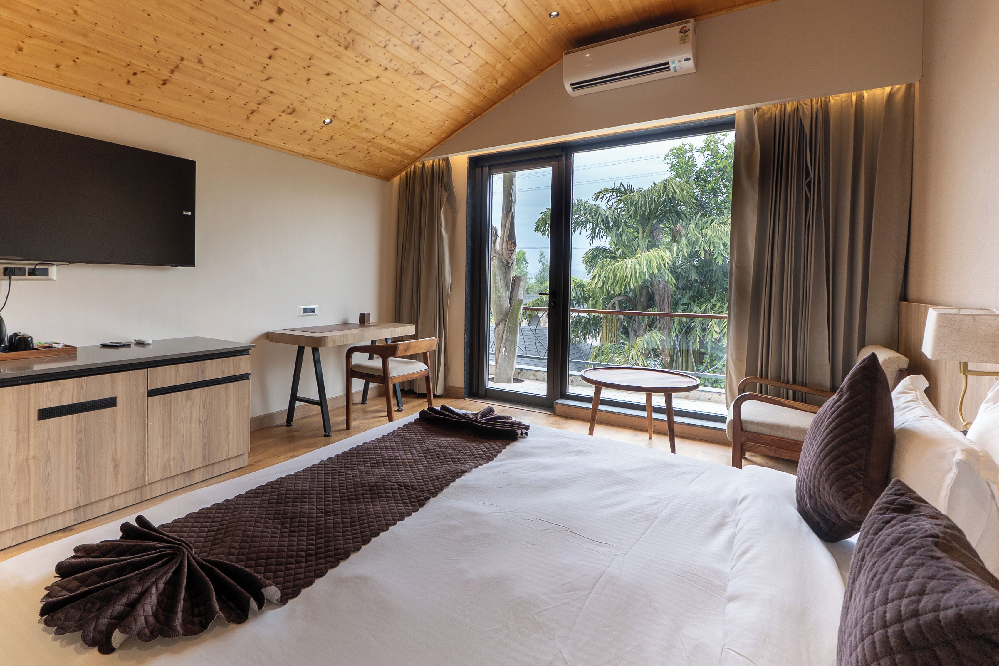A modern hotel room with a wooden ceiling, a flat-screen TV, a dining area, a balcony with curtains, and a large bed with pillows.