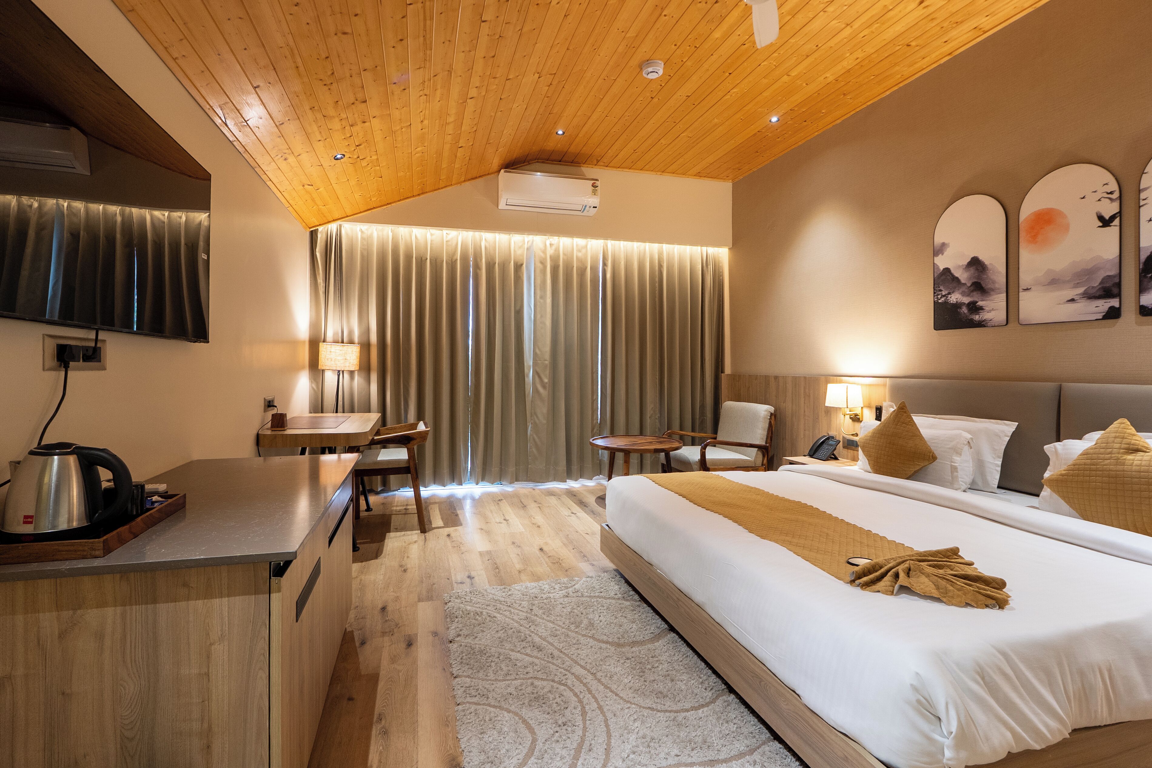 Habitación de hotel con techo de madera, cama con manta color beige, escritorio con hervidor, una silla, una mesita y televisor montado en la pared.