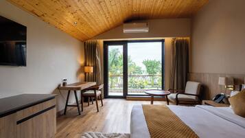 Habitación de hotel con techo de madera, cama, escritorio, silla, una mesita y balcón con vistas a zonas verdes.