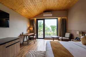 A hotel room with a wooden ceiling, a bed, a desk, a chair, a small table, and a balcony with a view of greenery. - juSTa Nature's Nest Resort (Rishikesh)