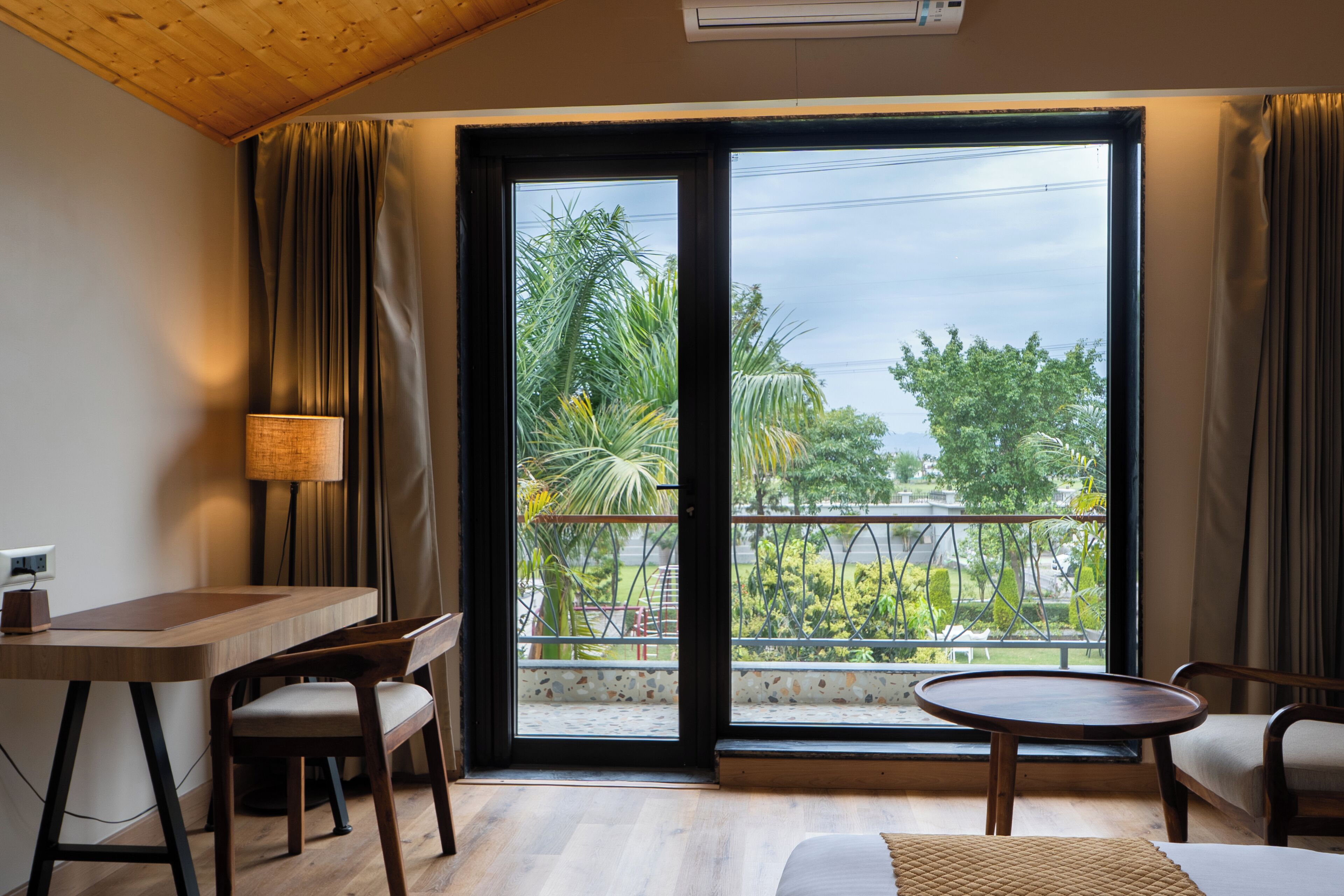Ein Zimmer mit Holzschreibtisch, zwei Stühlen, einem runden Tisch, einer Couch und einer großen Schiebeglasdoor, die Blick auf Grünfläche gewährt.