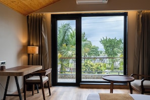 A room with a wooden desk, two chairs, a round table, a sofa, and a large sliding glass door with a view of greenery outside. - juSTa Nature's Nest Resort (Rishikesh)