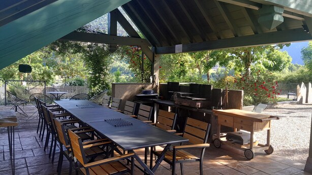 Une terrasse extérieure avec une longue table et des chaises, un barbecue et un espace abrité offrant une vue sur les arbres et les collines.