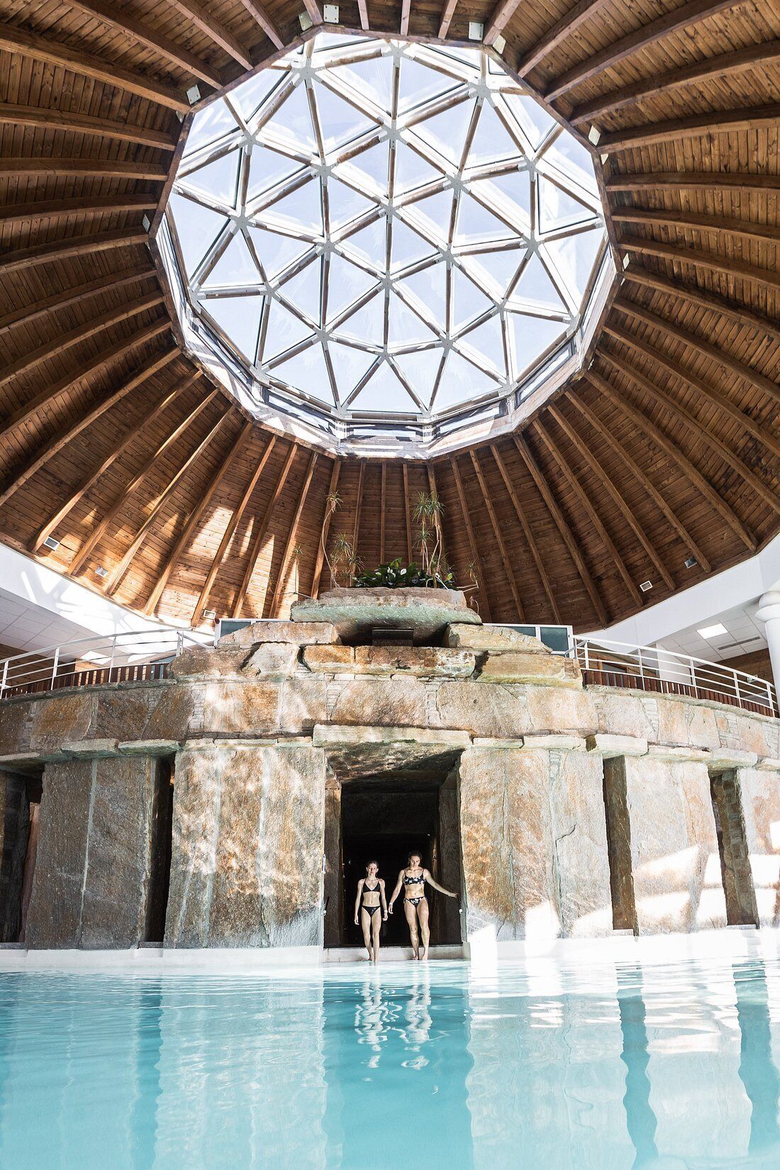 Une piscine intérieure dotée d’un grand plafond de verre aux formes géométriques et d’une architecture en pierre.