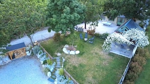Ein von einer Hecke umgebener Garten mit einer kleinen Gartenhütte, einem Baum mit weißen Blüten und einer Terrasse mit Gartenmöbeln.