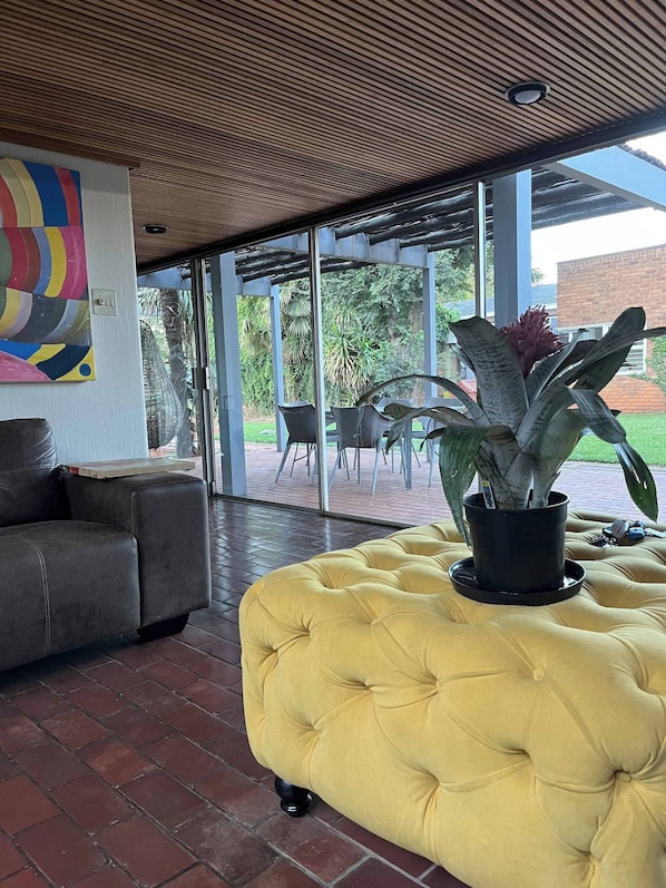 A modern interior with a yellow tufted ottoman, a grey sofa, and a large window overlooking a patio area with outdoor furniture.