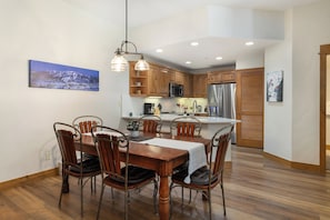 Un coin repas avec une table et des chaises en bois, une cuisine avec des armoires en bois et des appareils en acier inoxydable, et un tableau accroché au mur.