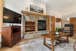A living room with a stone fireplace, a flat-screen TV, a wooden coffee table, and a leather armchair. - Steps from Slopes | Winter Shuttle | Pool & Hot Tub (Steamboat Springs)