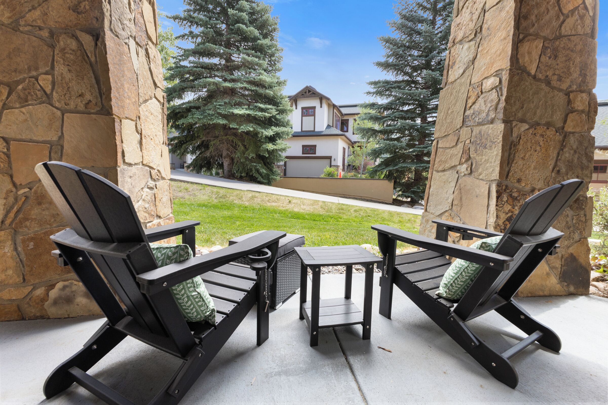 A patio with two Adirondack chairs and a small table, overlooking a lawn and a house.