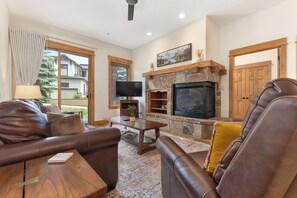 A living room with a stone fireplace, leather sofas, a television, and a view of a house through a window. - Steps from Slopes | Winter Shuttle | Pool & Hot Tub (Steamboat Springs)