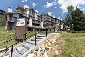 A multi-story building with a sign reading 'SKI TIME SQUARE,' a staircase with metal railings, and a grassy area with rocks. - Slopeside | Hot Tub | Bonus Den with Video Games (Steamboat Springs)