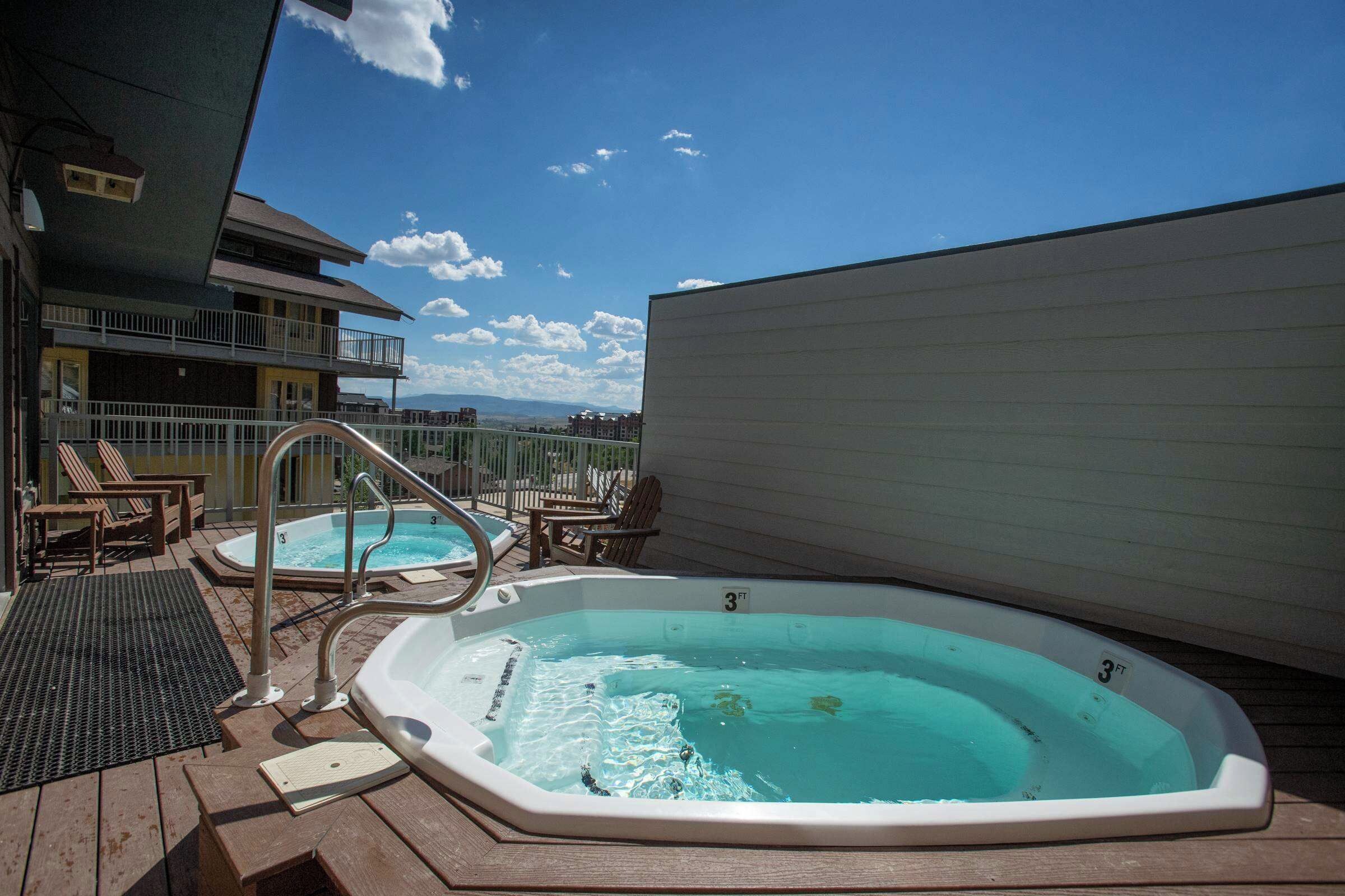 Un jacuzzi en una terraza de madera con vistas a un edificio y montañas al fondo.