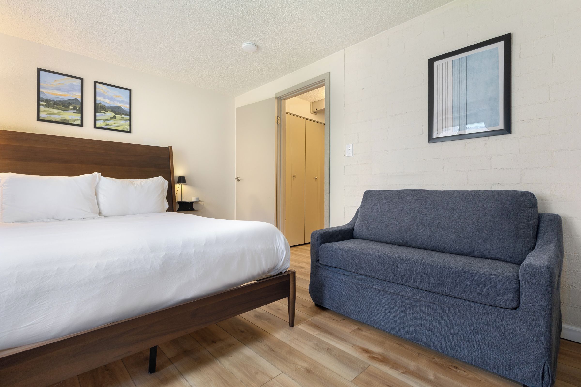 A hotel room with a bed, a grey sofa, wooden flooring, and two landscape paintings on the wall.