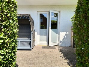 A white building with a sliding glass door, a covered outdoor seating area, and a paved patio. - Reiherweg, Apartment 2 - Beautiful 2-room vacation apartment with two terraces, quiet and (Sylt)