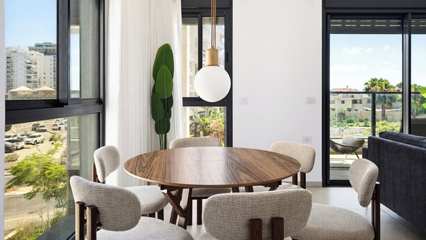 A modern dining area with a round table, six chairs, and a large window offering a city view. - Spacious 4BR Apt l Sunny Balcony & Mamad (Bat Yam)