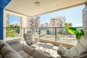 A balcony with a sofa, chair, and small table, overlooking a cityscape with buildings and a road. - Spacious 4BR Apt l Sunny Balcony & Mamad (Rishon LeTsiyon)