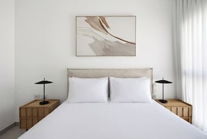 Uma cama bem arrumada com duas mesas de cabeceira, cada uma com uma luminária, em um quarto simples com uma parede lisa e uma única obra de arte.