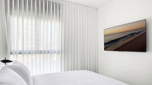 A hotel room with a bed, white curtains, a wall-mounted TV, and a window with blinds. - Spacious 4BR Apt l Sunny Balcony & Mamad (Bat Yam)