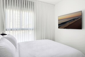 A hotel room with a bed, white curtains, a wall-mounted TV, and a window with blinds. - Spacious 4BR Apt l Sunny Balcony & Mamad (Bat Yam)