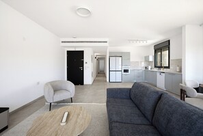 Uma sala de estar moderna com um sofá cinzento, uma mesa de centro redonda e uma poltrona cinzenta. Uma cozinha com eletrodomésticos e armários brancos é visível ao fundo.