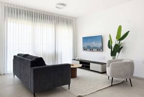 A modern living room with a sofa, armchair, coffee table, and a TV on a stand. - Spacious 4BR Apt l Sunny Balcony & Mamad (Bat Yam)