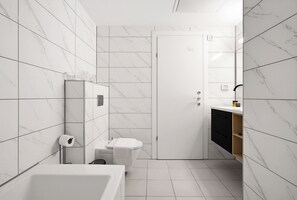 Um banheiro moderno com azulejos de mármore branco, uma banheira branca, vaso sanitário e um espelho sobre um móvel de madeira.