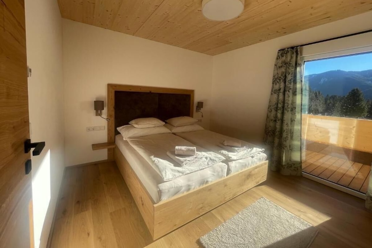A bedroom with a wooden ceiling, a bed with white linens, a nightstand, a window with a view of mountains, and a wooden floor.