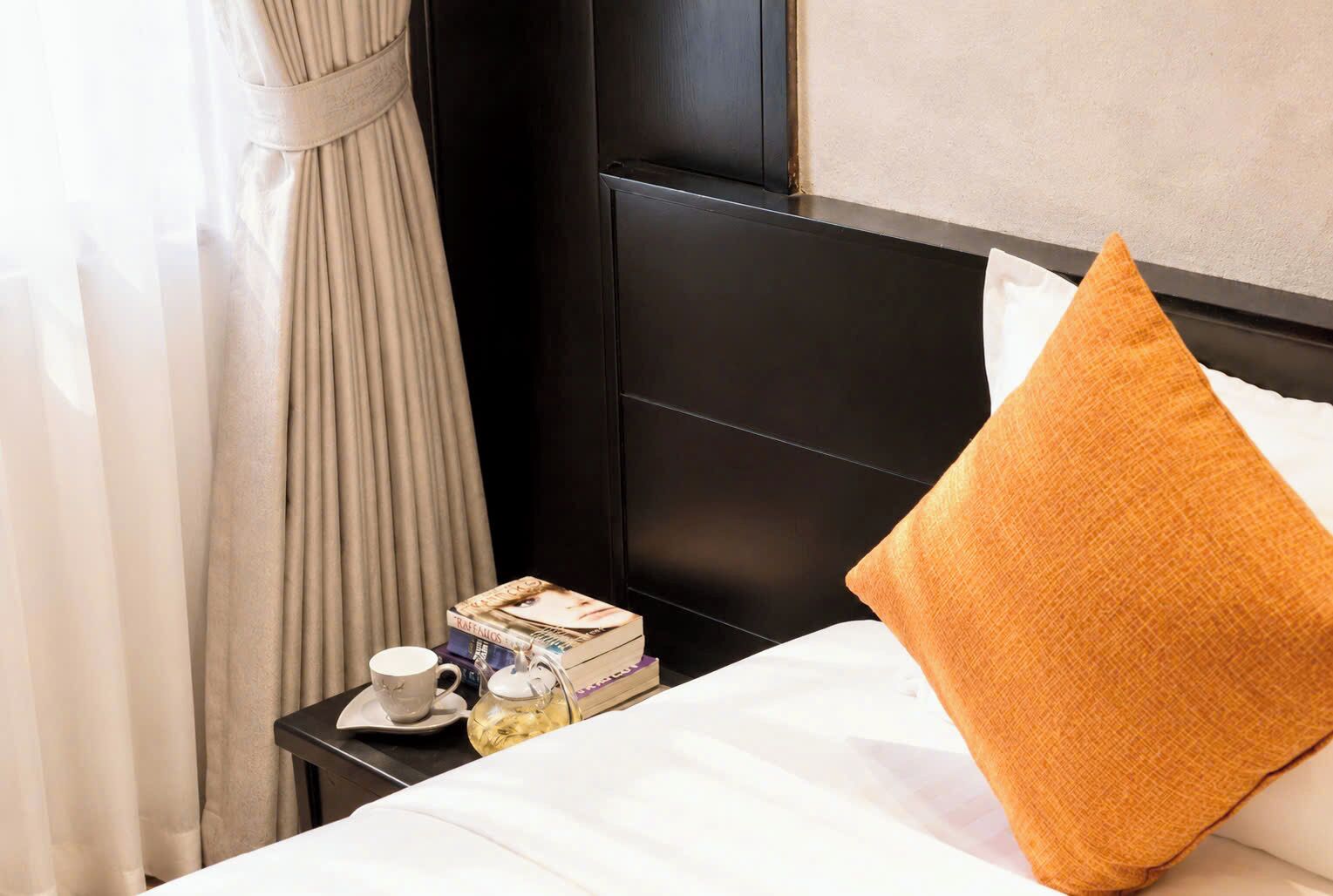 Una cama bien hecha con una colcha blanca y una almohada naranja, una mesita de noche pequeña con una taza, una tetera y una pila de libros.