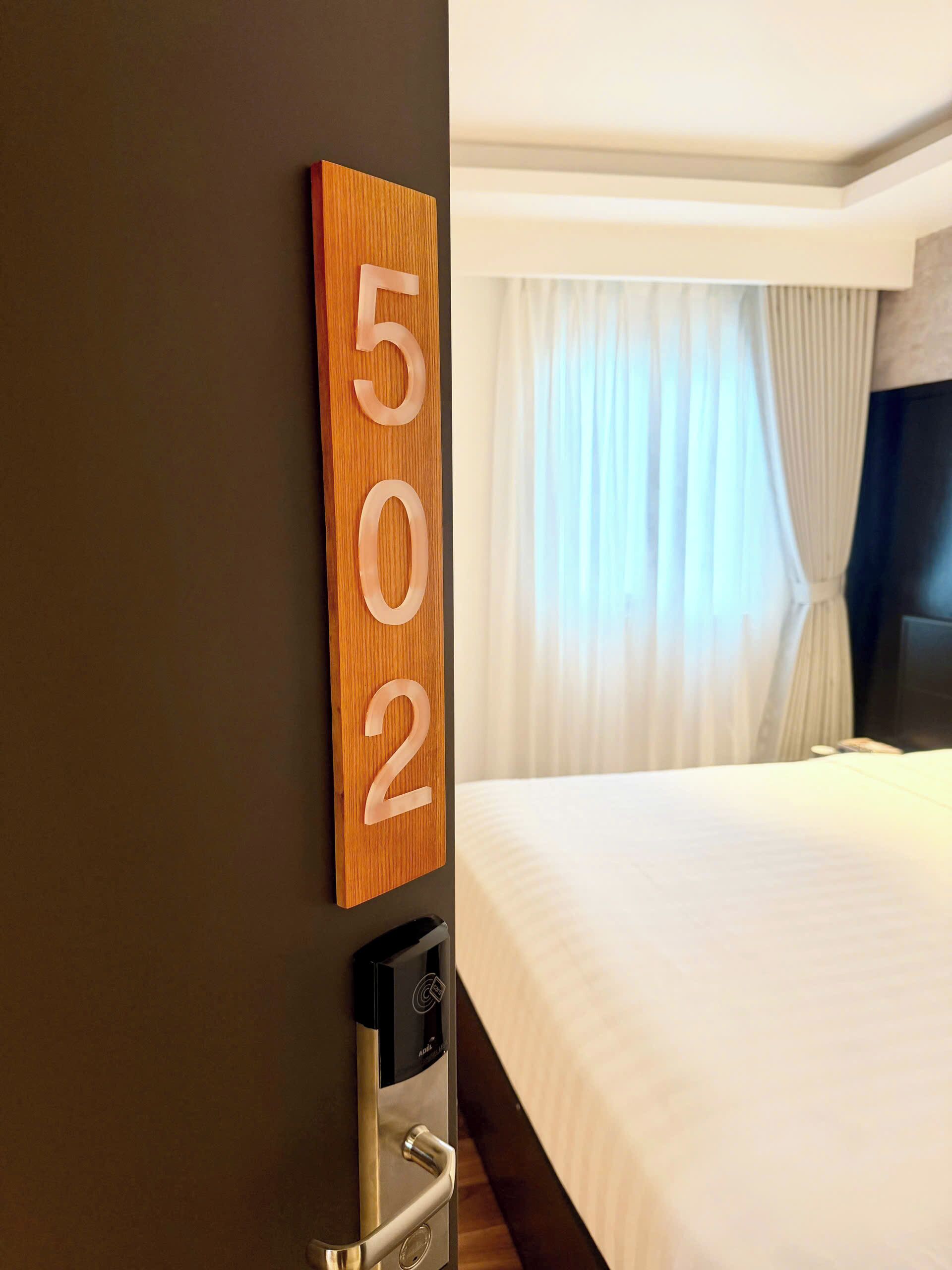 A hotel room with a wooden number sign indicating room 502, a bed with white bedding, and a door with a keycard reader.