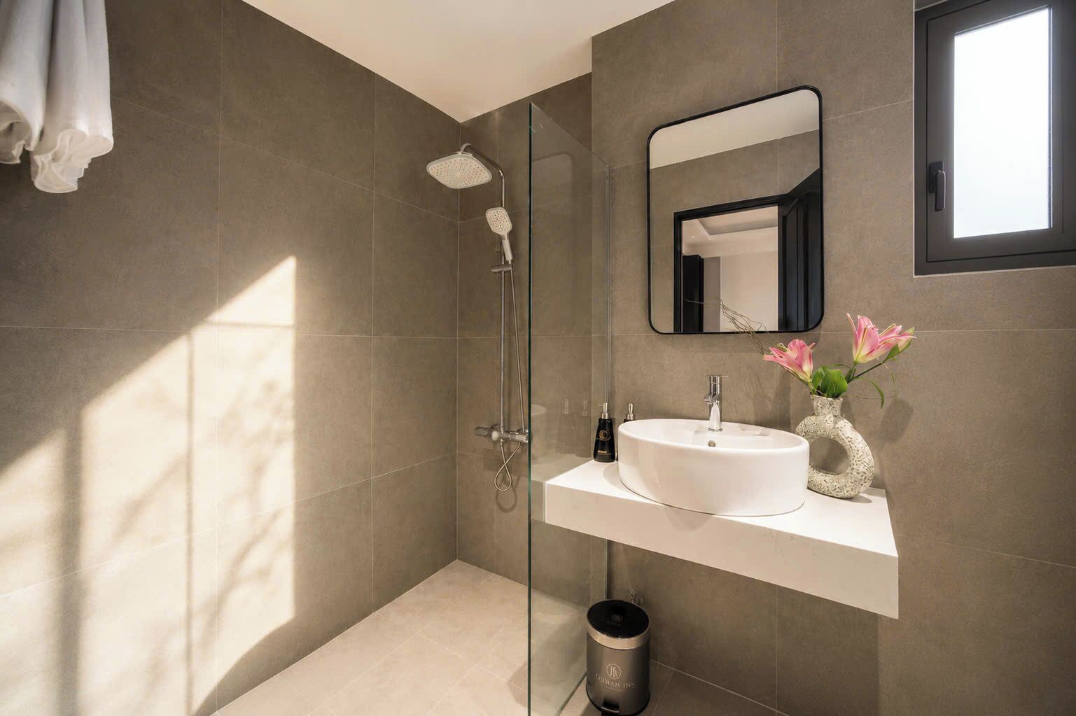 Un baño moderno con ducha envuelta en vidrio, un lavabo blanco con un florero y un espejo grande.