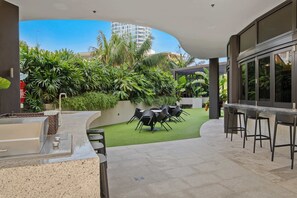 A patio area with a grill, outdoor seating, and a view of a building and greenery. - 『CASSA VUE』- 3-BR Coastal Living Apartments (Broadbeach)