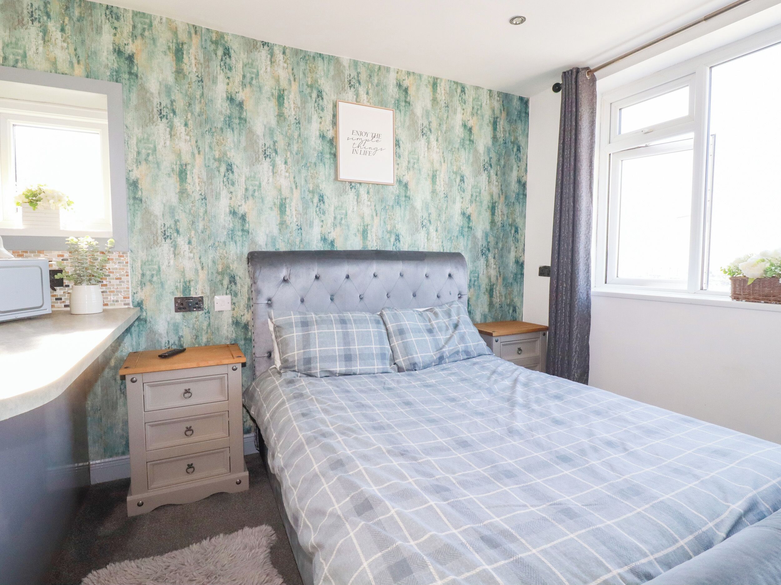 A bedroom with a bed, a bedside table, a dresser, a wall clock, a framed quote, and a window with curtains.