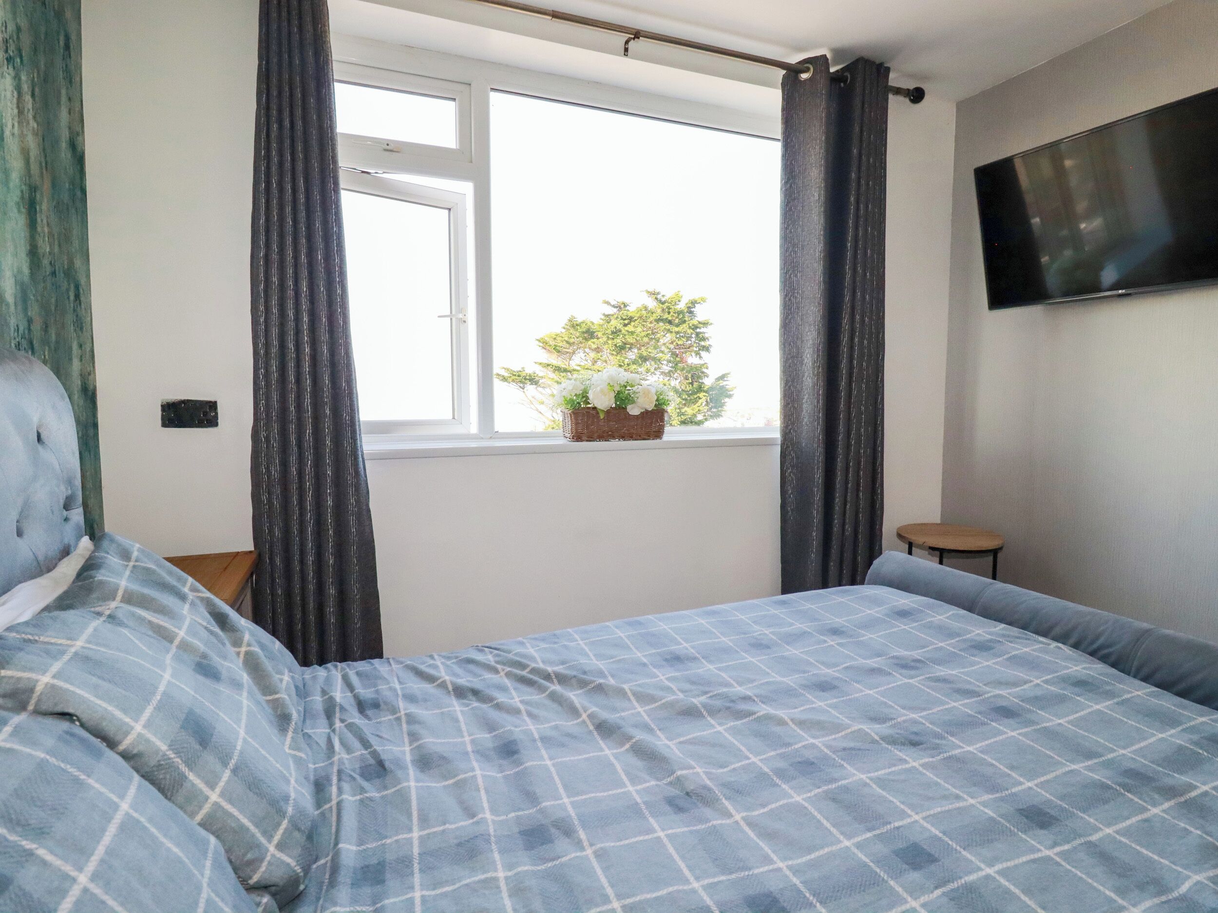 Ein Schlafzimmer mit einem Bett, einem Fenster mit Vorhängen, einem kleinen Tisch und einem an der Wand montierten Fernseher.