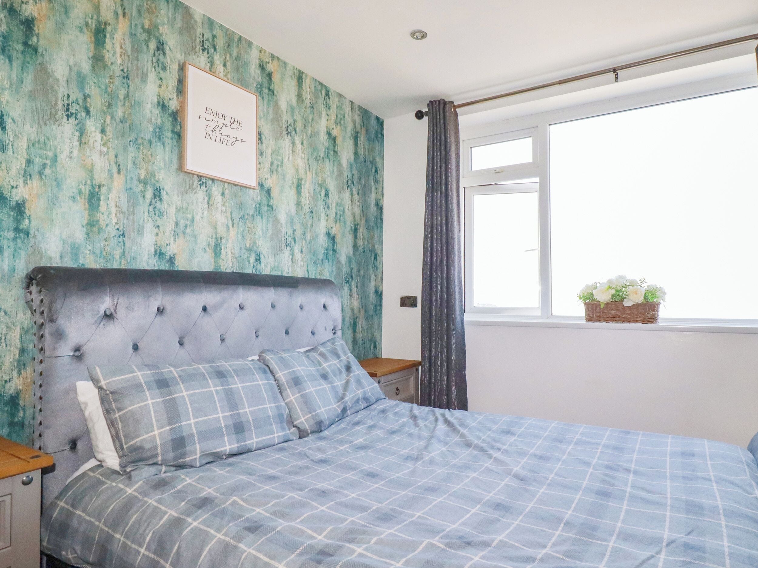 A bedroom with a tufted headboard, a bed with a blue checkered bedspread, a framed inspirational quote on the wall, a window with a view of the outside, and a small plant on the windowsill.