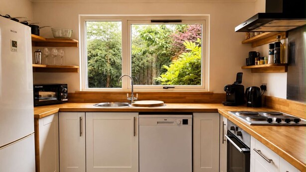 A modern kitchen with a dishwasher, stove, and a window overlooking greenery. - Ivy & Ink Cottage - Inverness (Inverness)