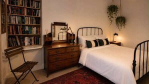 A bedroom with a wooden bookshelf, a mirror, a bed with white bedding, a red patterned rug, and hanging plants. - Ivy & Ink Cottage - Inverness (Inverness)
