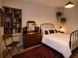 A bedroom with a wooden bookshelf, a mirror, a bed with white bedding, a red patterned rug, and hanging plants. - Ivy & Ink Cottage - Inverness (Inverness)