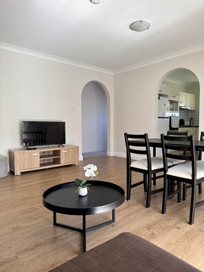 A modern living room with a TV, dining table, chairs, and a coffee table with a flower. - A beautiful room in Clayfield, Brisbane - 10 min to the airport! (Clayfield)