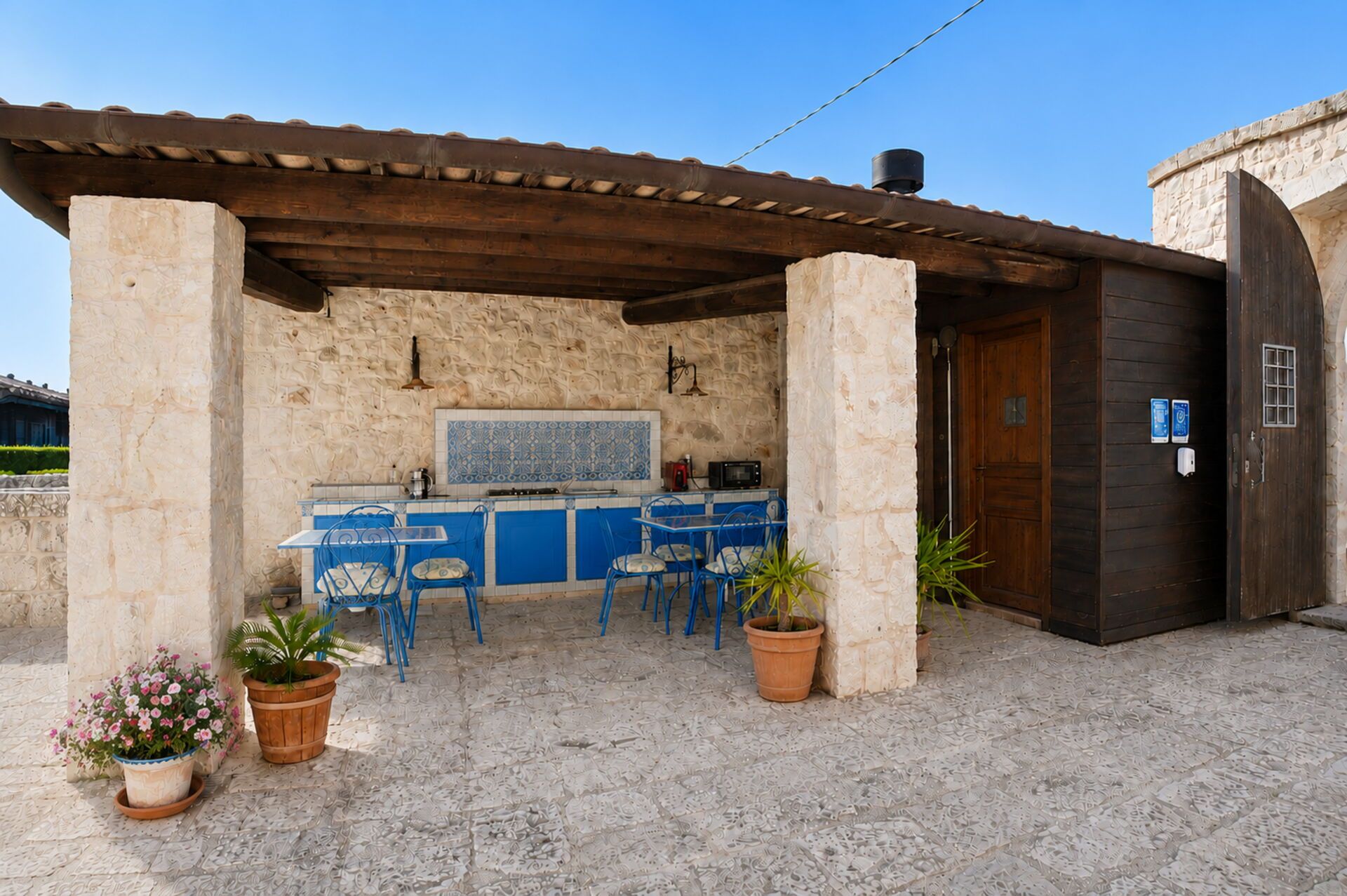 A covered outdoor kitchen area with a stone wall, blue chairs, and potted plants.